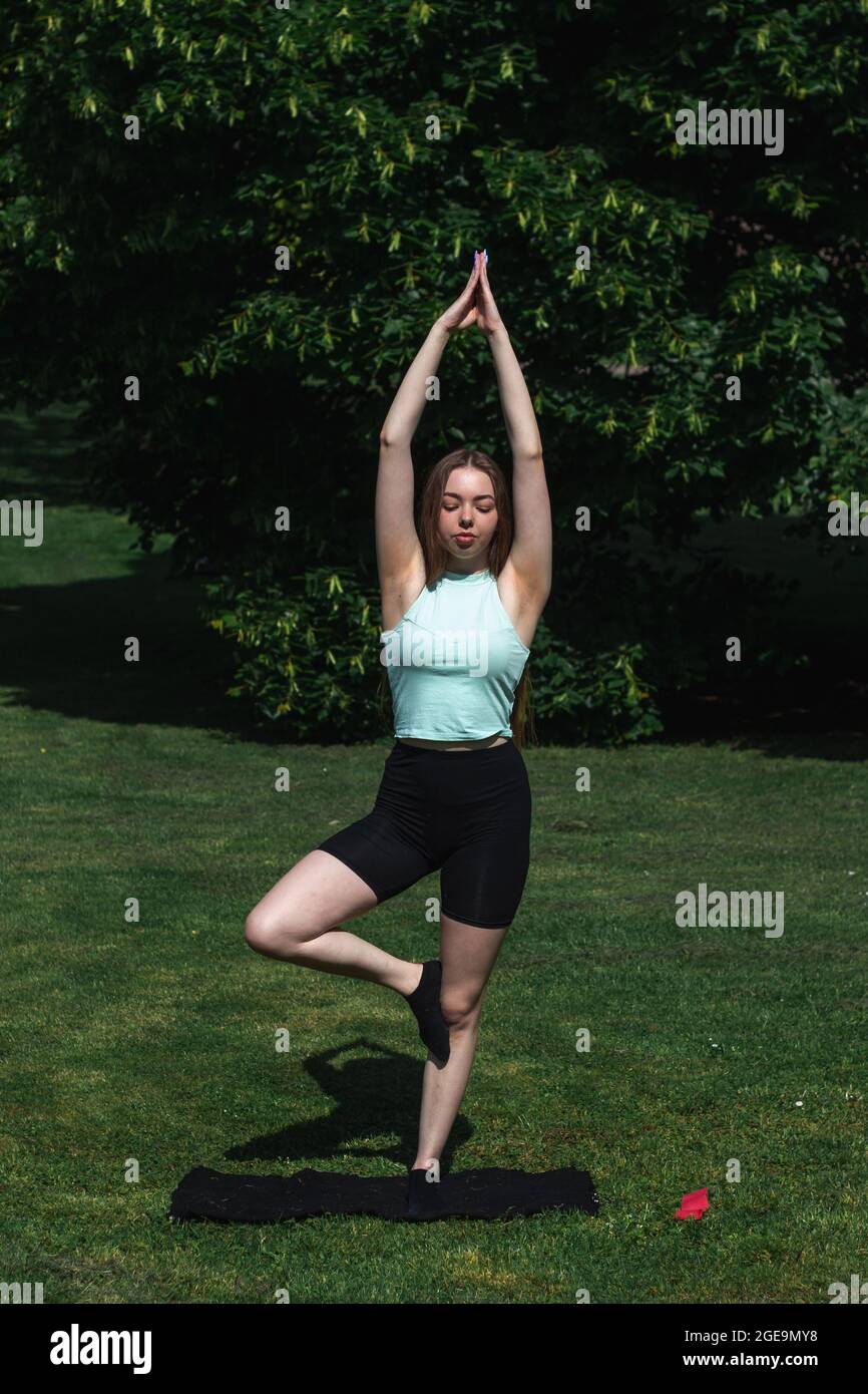 Giovane caucasica dai capelli lunghi in abbigliamento sportivo sta facendo yoga nel parco pubblico. Allenamento all'aperto. Lo yoga pone Foto Stock