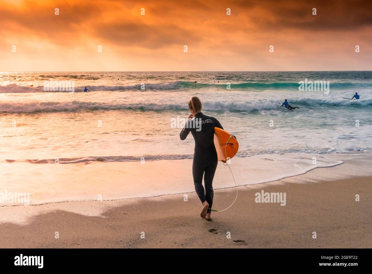 Una surfista femminile che porta la sua tavola da surf e che cammina in mare in tarda serata alla luce del sole a Fistral Beach a Newquay in Cornovaglia. Foto Stock