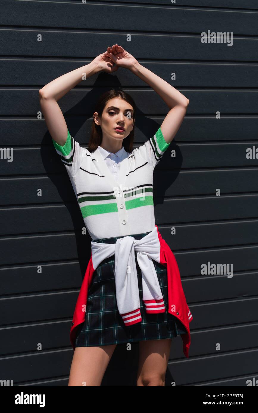 giovane donna in polo a righe in posa vicino a parete grigia con mani rialzate Foto Stock