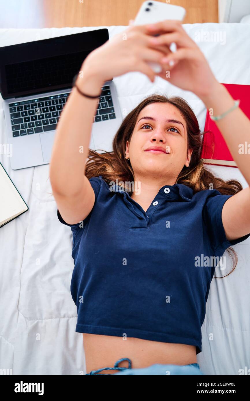 Dall'alto della giovane studentessa positiva che si trova a letto e prende selfie sullo smartphone mentre si è pausa durante gli studi remoti online a casa Foto Stock