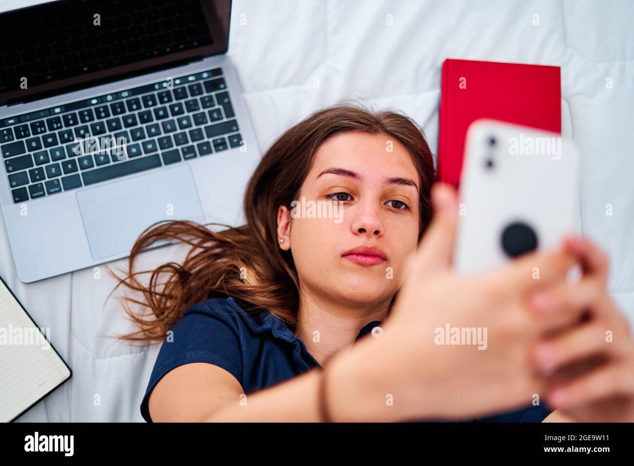 Dall'alto della giovane studentessa positiva che si trova a letto e prende selfie sullo smartphone mentre si è pausa durante gli studi remoti online a casa Foto Stock