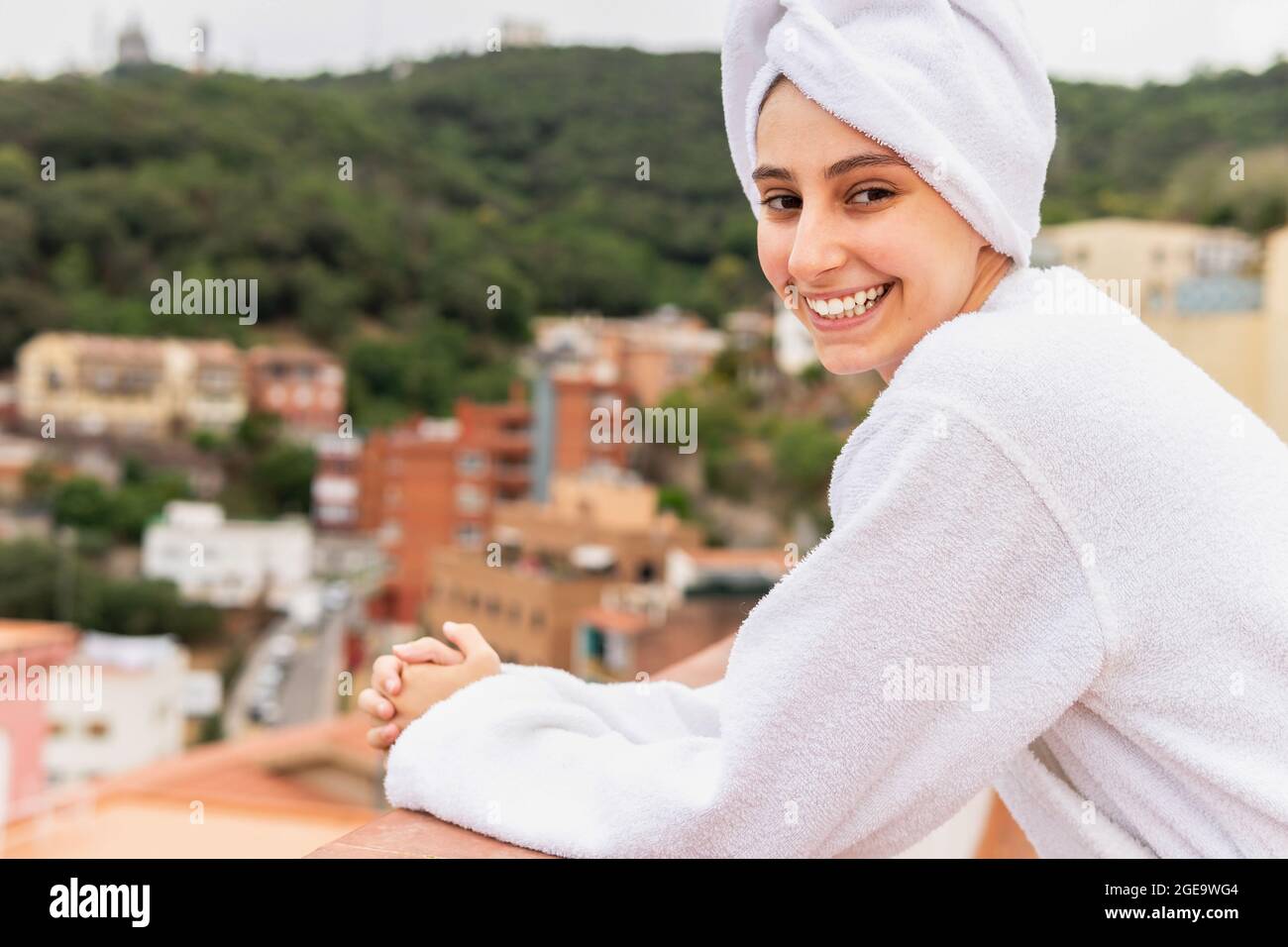 Giovane donna ottimista in accappatoio e asciugamano sorridendo e guardando la macchina fotografica mentre si rilassa sul balcone durante la routine di cura della pelle nel fine settimana Foto Stock