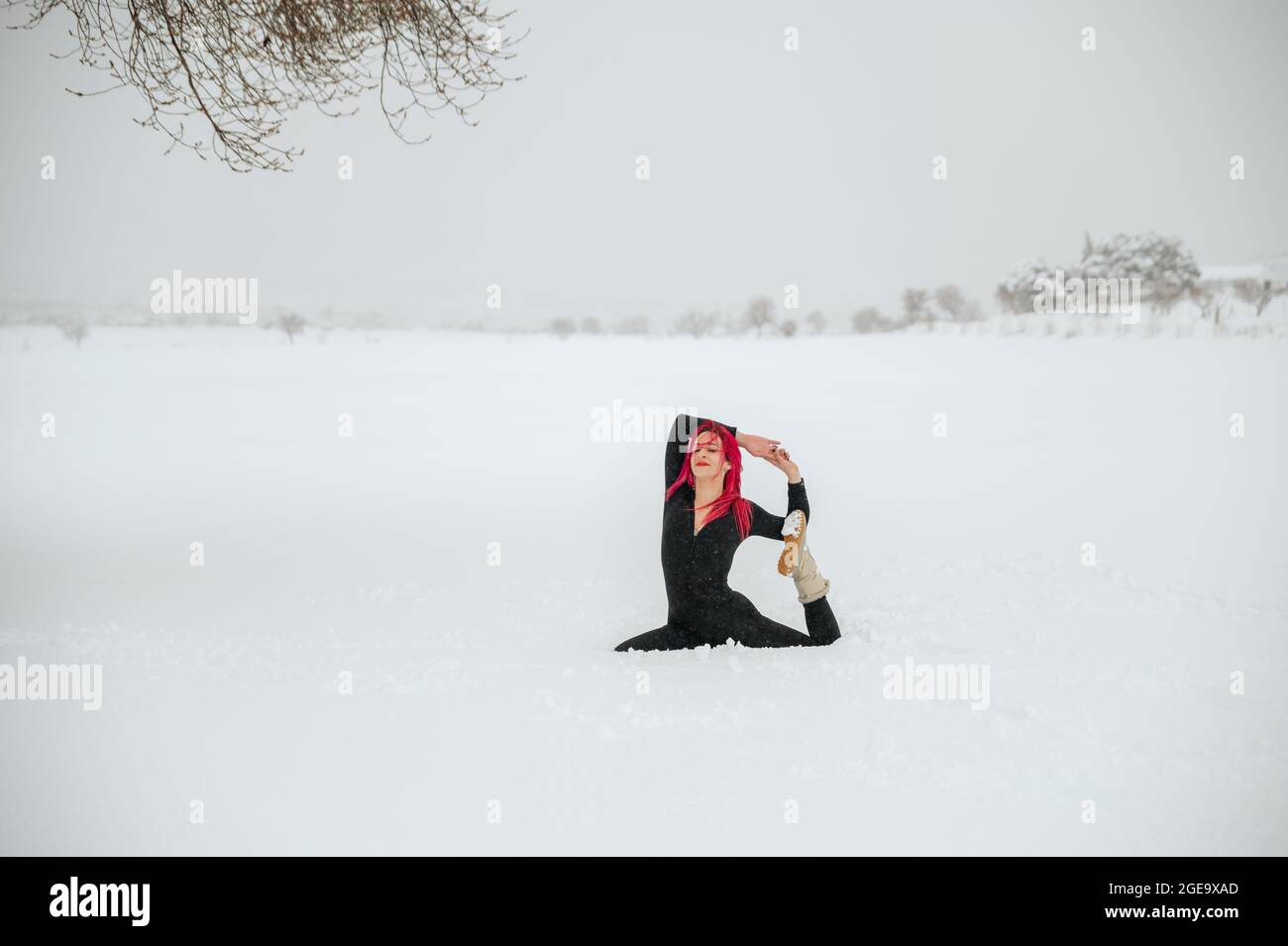 Donna serena con capelli rosa seduta in Eka Pada Raja Kapotasana su campo invernale nevoso e praticare yoga Foto Stock