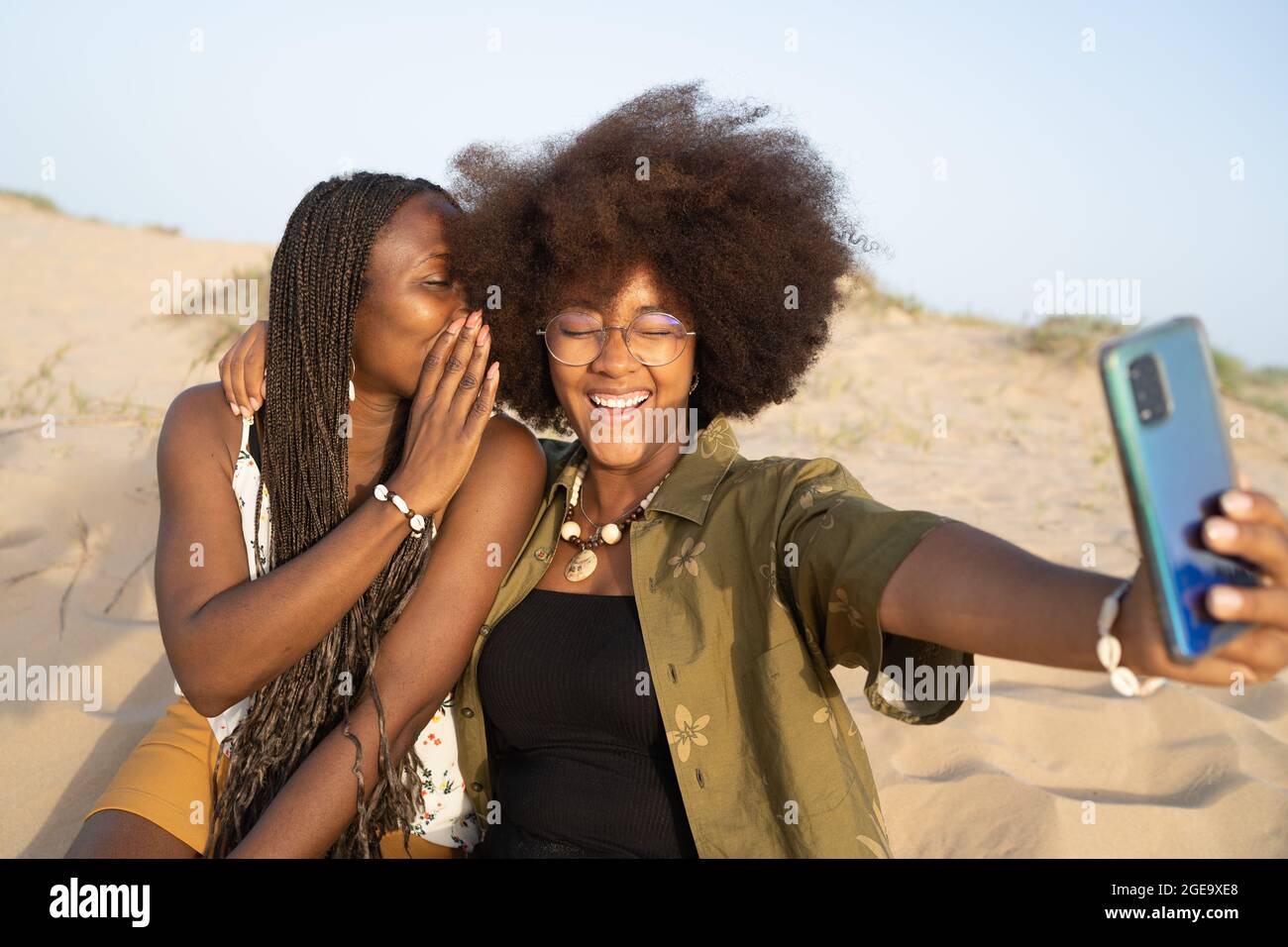 Allegra giovane afroamericana femmina amici sorridente mentre prendendo selfie su smartphone durante il fine settimana estivo sulla spiaggia sabbiosa Foto Stock