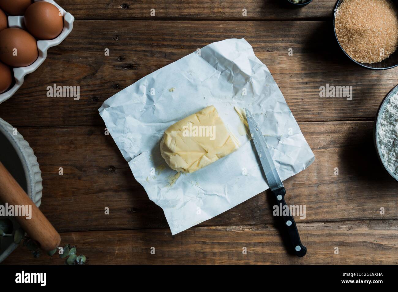 Coltello pronto a tagliare il pezzo di burro su una preparazione di pasticceria su tavola di legno vicino alle uova e zucchero di canna Foto Stock