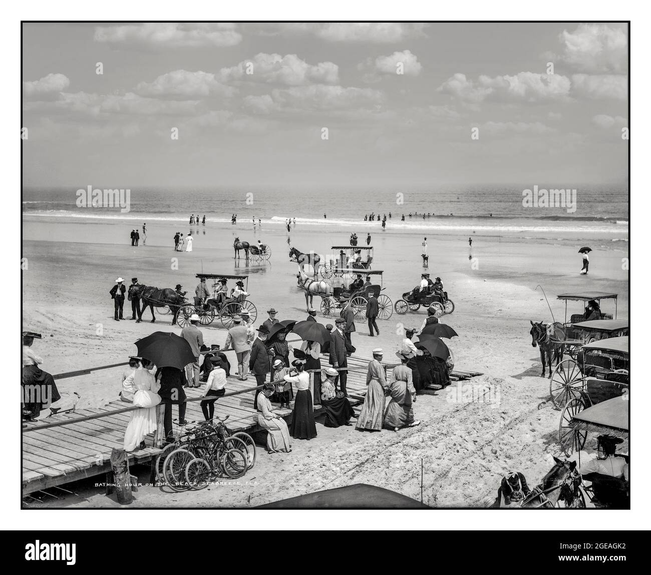 Spiaggia vittoriana in Florida, carrozze da spiaggia trainate da turisti e cavalli e un'auto da corsa Oldsmobile del 1904, godendosi il sole al resort sulla costa della Florida con ombrelloni e abbigliamento formale anni '1900 che fa il bagno e visita sulla spiaggia di Daytona, Florida USA 1904 Foto Stock
