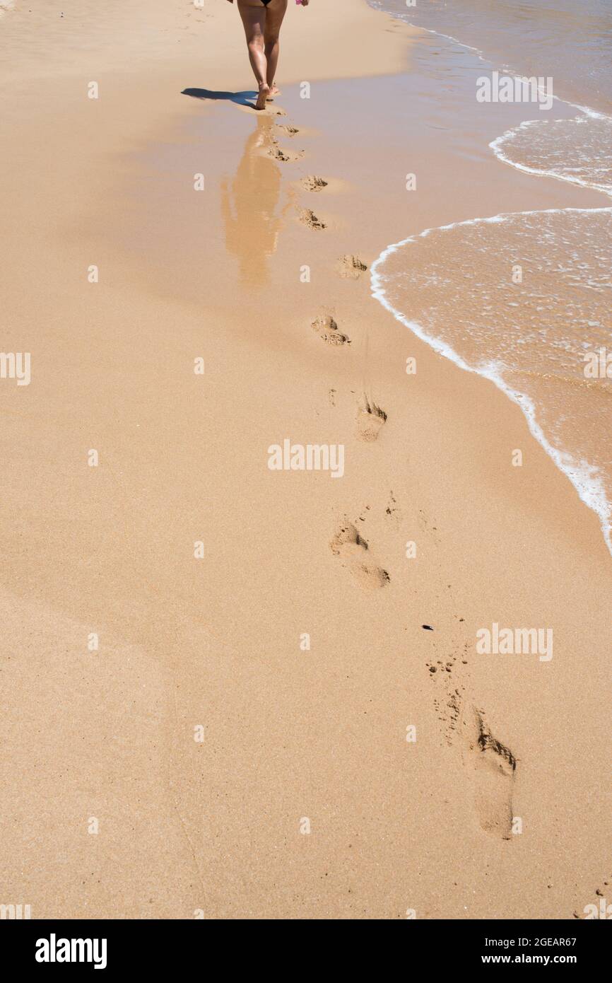 Donna caucasica vista dalla sua schiena camminando su una spiaggia vuota. Impronte nella sabbia. Il sistema circolatorio è migliorato dall'esercitazione aerobica, come camminare Foto Stock