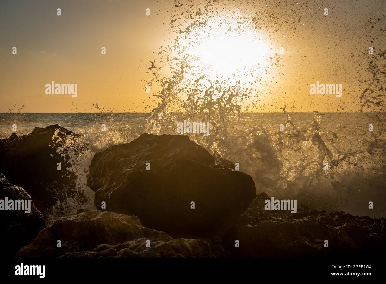 Onde che si infrangono sulle rocce della spiaggia con il sole che tramonta sotto l'orizzonte. Foto Stock