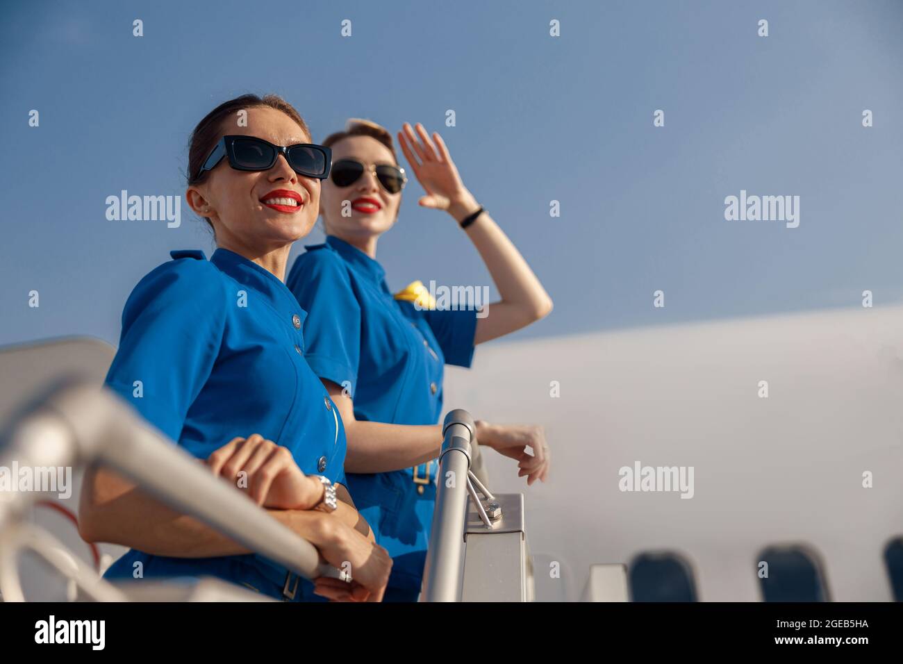 Ritratto di due allegre passerette d'aria in uniforme blu e occhiali da sole che sorridono, in piedi insieme su un airstair in una giornata di sole Foto Stock