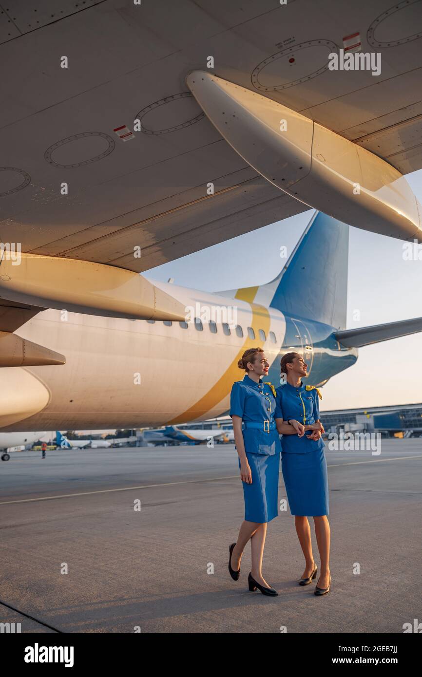 Foto a lunghezza intera di due stewardesses d'aria in uniforme blu che sorridono, in piedi di fronte a un grande aereo passeggeri in aeroporto al tramonto Foto Stock