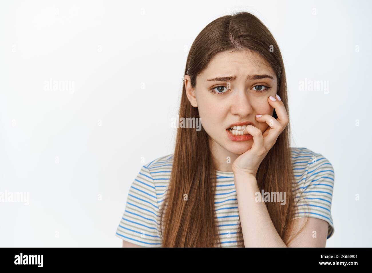 Ritratto di una ragazza spaventata che le mordicchiava il dito, guardando ansioso e preoccupato per la macchina fotografica, in piedi in t-shirt su sfondo bianco Foto Stock