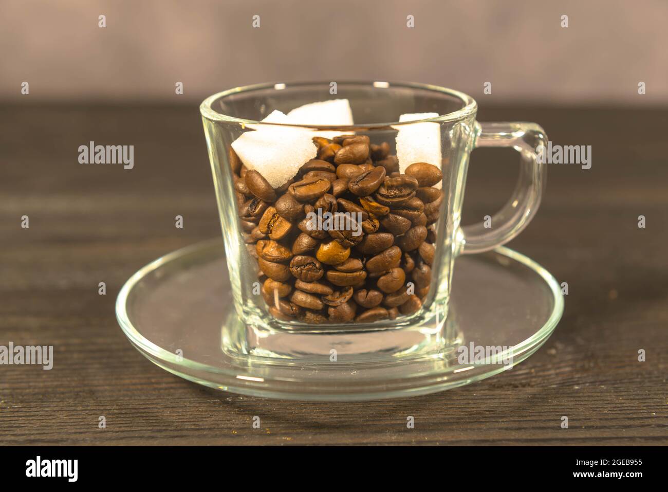 Una tazza di vetro piena di chicchi di caffè marroni. Si trova su una superficie scura con il piattino. Ci sono due cubetti di zucchero bianco sulla parte superiore dei chicchi di caffè. Foto Stock