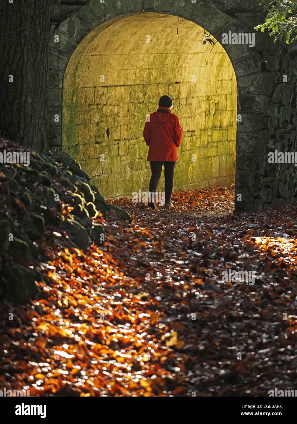 Donna lone con cappello e cappotto rosso che cammina nel tunnel arcuato di pietra pieno di luce verde a Cumbria, Inghilterra, Regno Unito Foto Stock