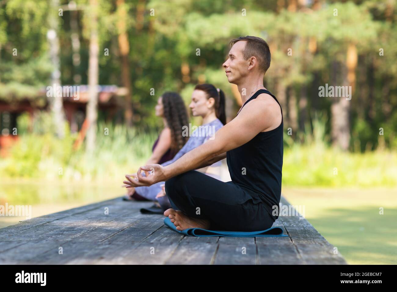 Tre persone, un uomo e due donne, sono impegnate nella meditazione seduta su un tappetino in posizione loto su un ponte di legno nel parco in estate soleggiata Foto Stock