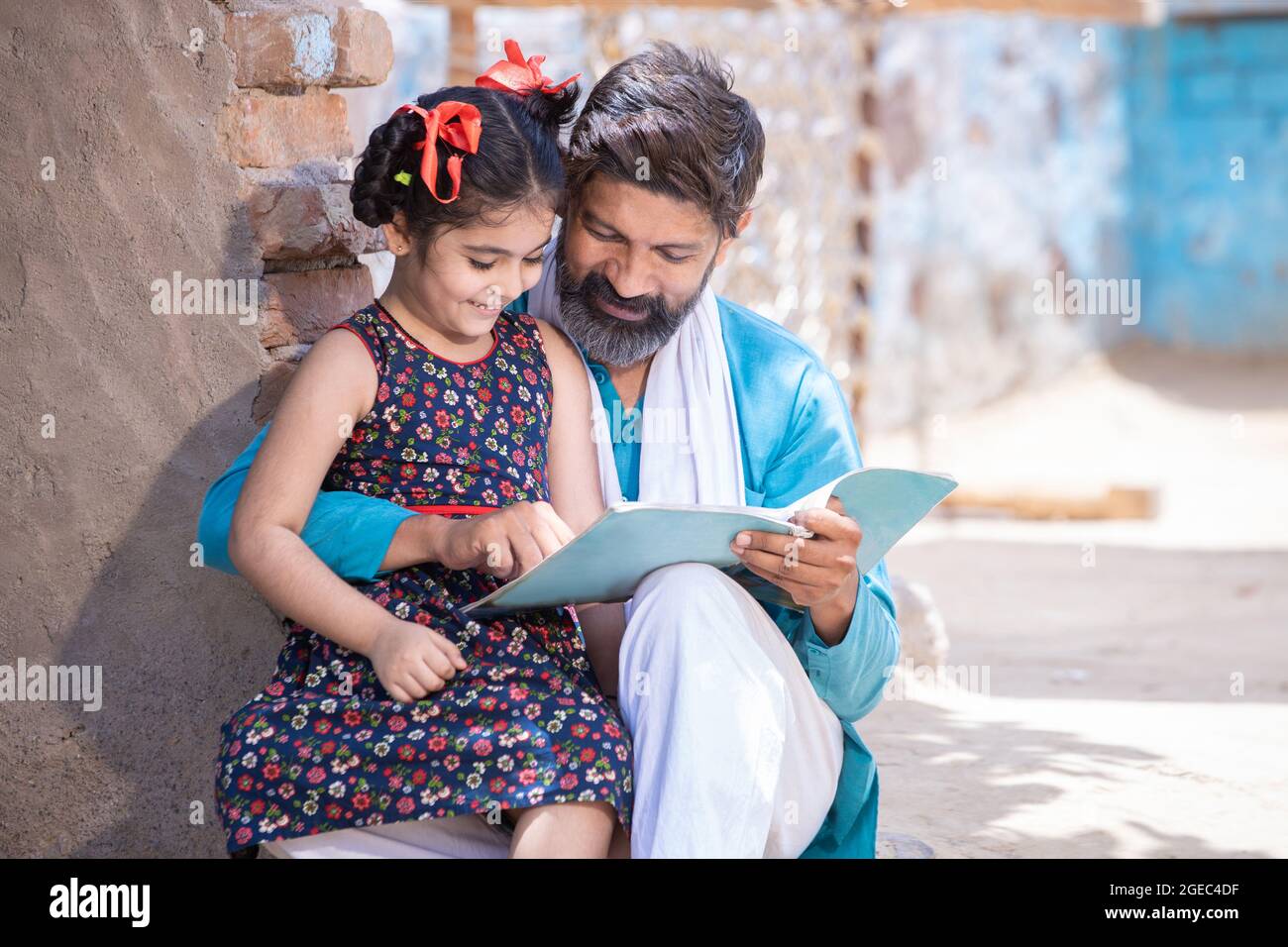 Felice padre indiano rurale aiutando sua figlia negli studi, adorabile bambina del villaggio bambino tenendo notebook studiare fuori casa. L'uomo insegna e. Foto Stock