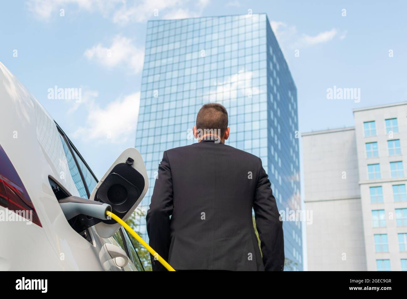 Uomo d'affari che collega il caricabatterie in auto elettrica e che va a lavorare nel business center - grattacielo Foto Stock