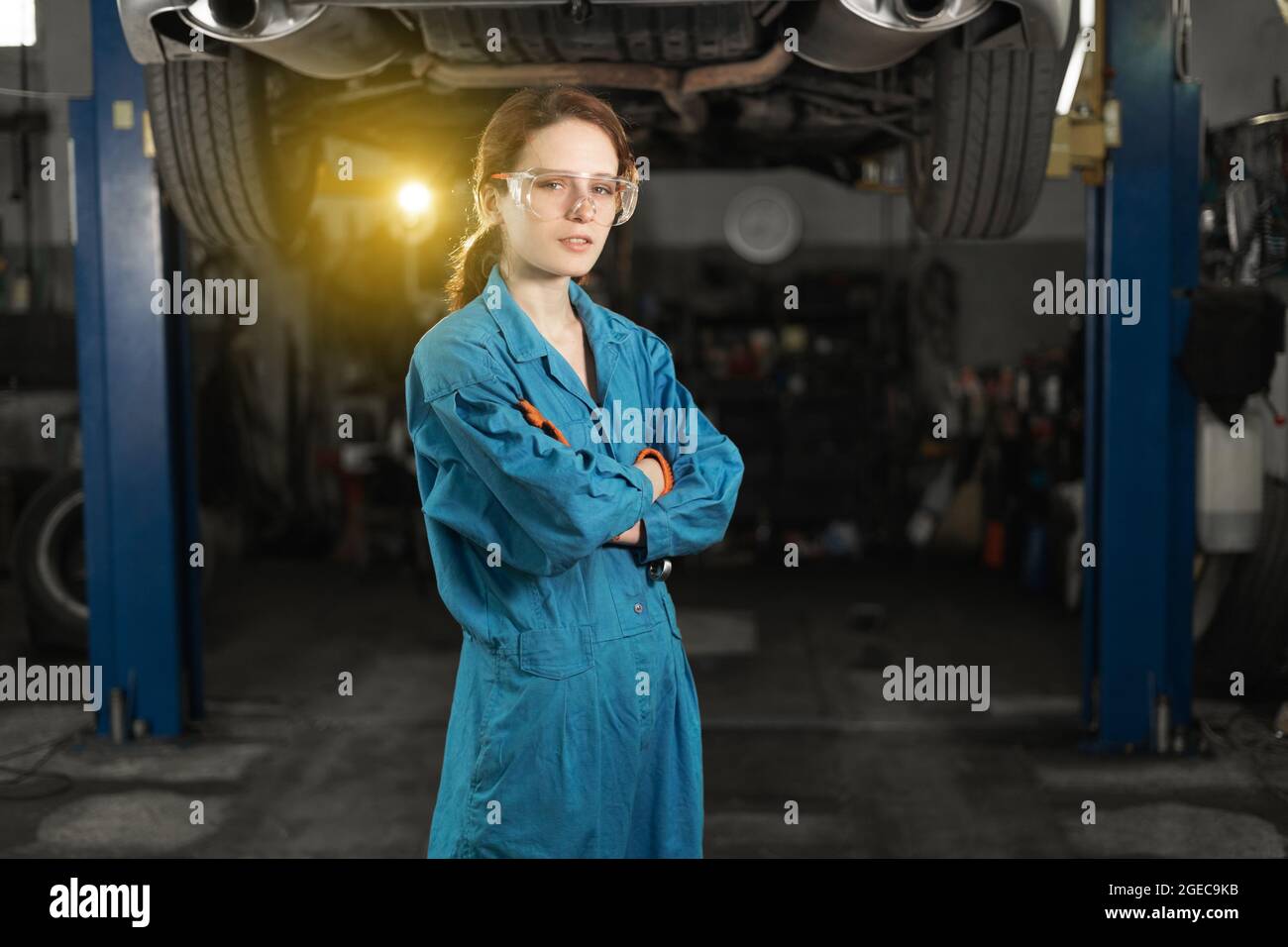 Una giovane donna in abiti da lavoro, un apprendista si trova accanto all'auto ed è orgogliosa e felice nel garage con occhiali protettivi. Spazio di copia. Foto Stock