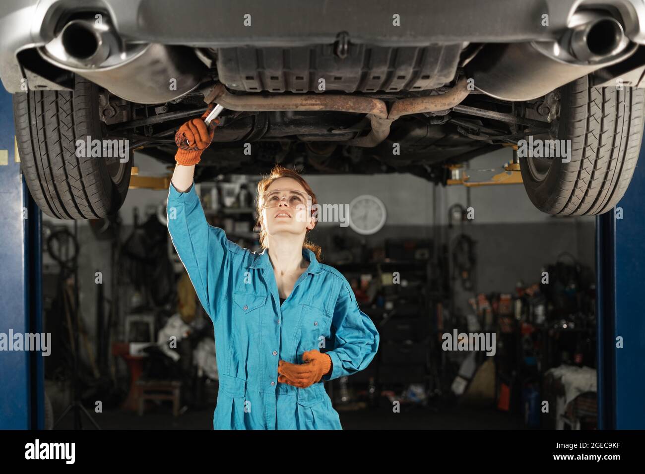 Bellissimo ritratto di una macchina professionale femminile che lavora sotto un veicolo su un ascensore in servizio. Utilizza una chiave. Lo specialista indossa protezioni Foto Stock