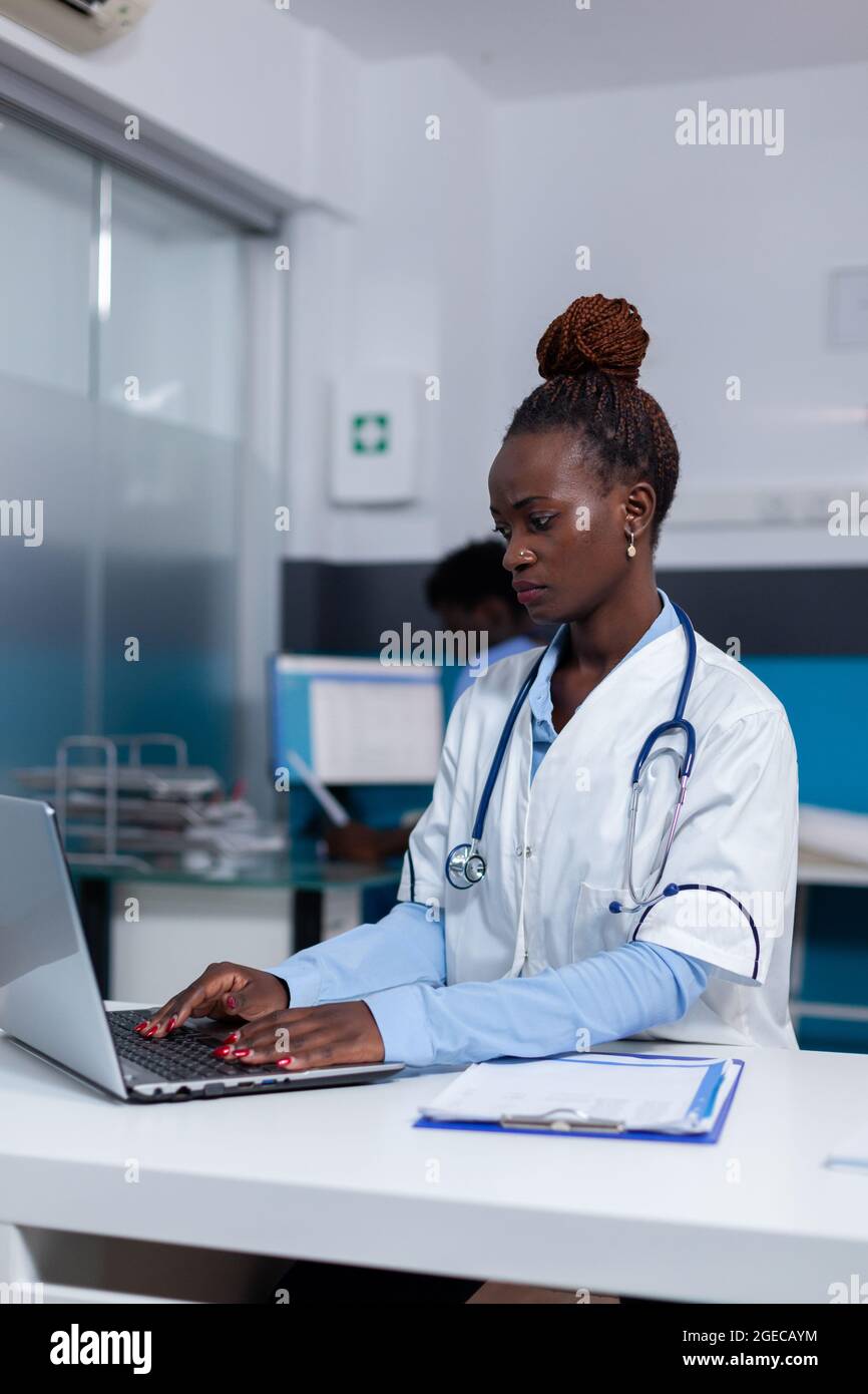 Persona afroamericana con lavoro medico utilizzando il computer portatile  alla scrivania in ufficio medico. Medico nero seduto con tecnologia e attrezzature  sanitarie mentre l'infermiere guarda il computer in background Foto stock -