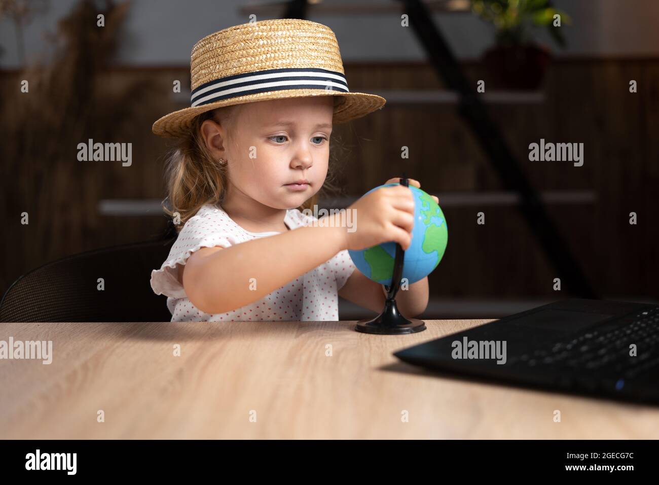 Una studentessa caucasica vestita con un cappello che tiene in mano un globo si siede a casa a un tavolo con un computer portatile. Foto Stock