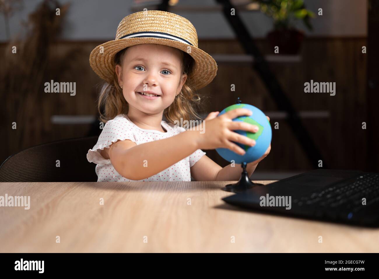 La ragazza carina caucasica in un cappello si siede sola la sera ad un tavolo con un laptop che tiene un globo nelle sue mani. Foto Stock