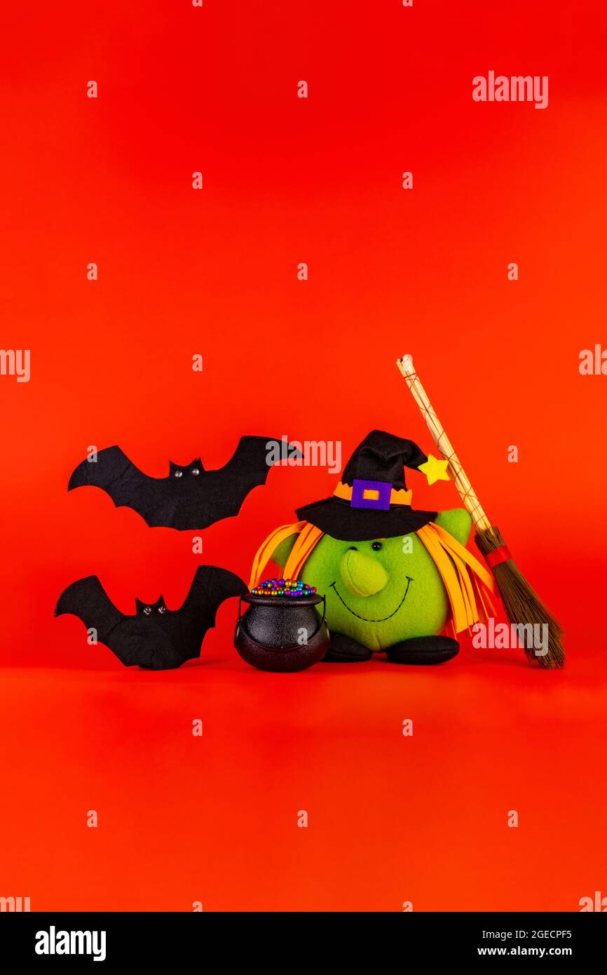 Bambola di strega verde, con il suo cavoldro e il broomstick insieme a due graziosi pipistrelli su sfondo rosso arancione. Concetto di tema di Halloween. Foto Stock
