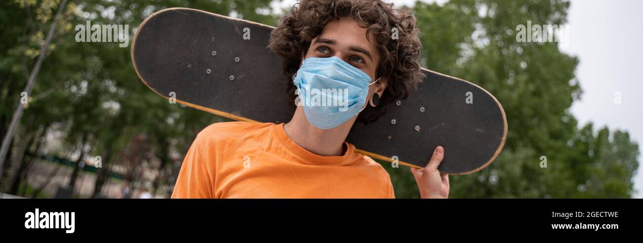 uomo curly in maschera medica tenendo skateboard dietro il collo, banner Foto Stock