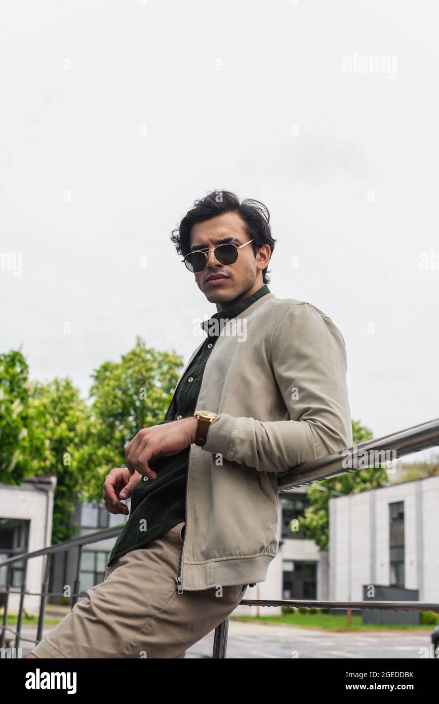 giovane uomo in eleganti occhiali da sole e giacca bombardiera in posa all'esterno Foto Stock