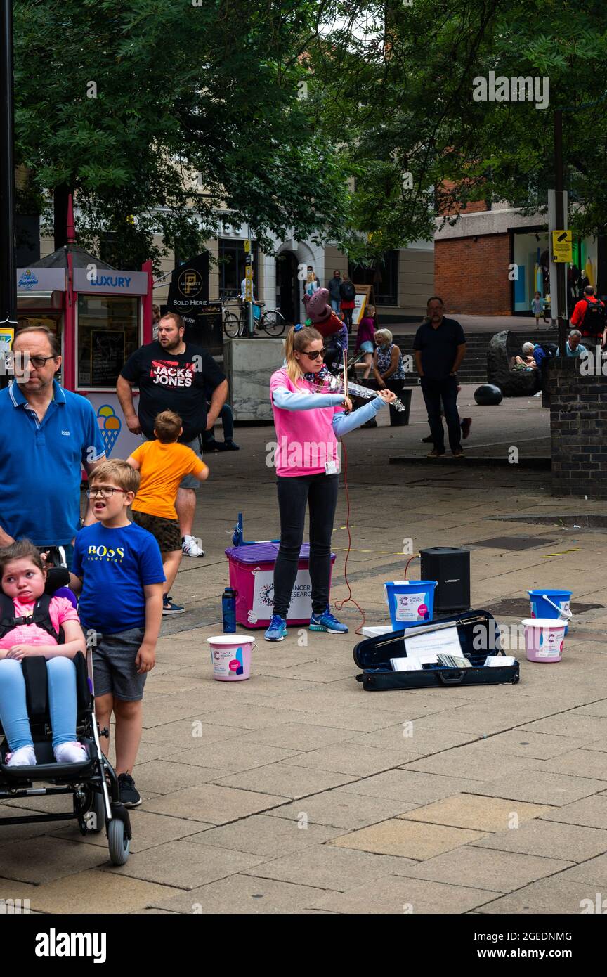 Young Lady giocando un violino elettrico nel mezzo del centro di Norwich con ricerca sul cancro UK secchi per le persone a mettere denaro in Foto Stock