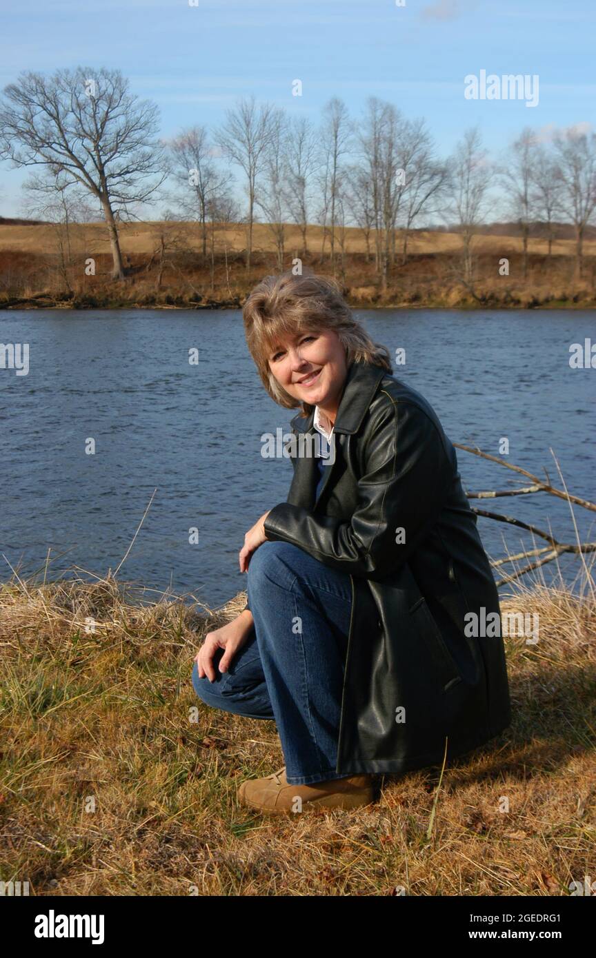 Donna sorridente e matura si inginocchia oltre al fiume in una zona rurale del Tennessee. Indossa un cappotto di pelle nera e jeans. Foto Stock