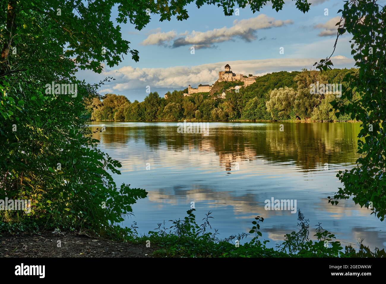 Castello sopra il fiume Vah. Vista della sponda destra. Incorniciato da foglie di albero. Riflesso di belle nuvole sulla superficie dell'acqua. Trencin, Slovacchia. Foto Stock