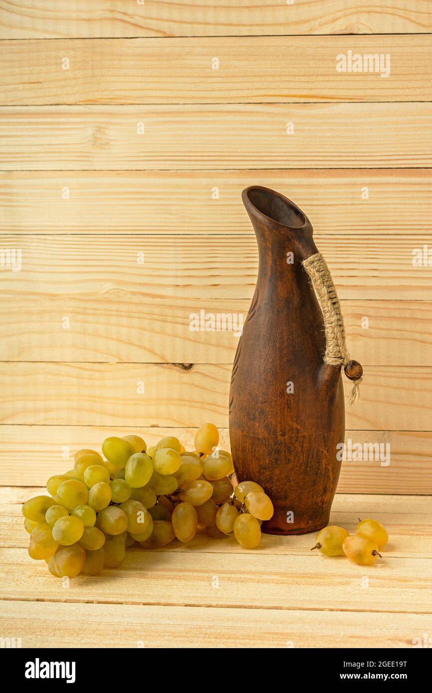 Caraffa d'uva su sfondo naturale in legno. Stile rustico retrò. Caraffa in argilla antica con vino. Uve verdi succose mature della vendemmia autunnale. Il concetto o Foto Stock