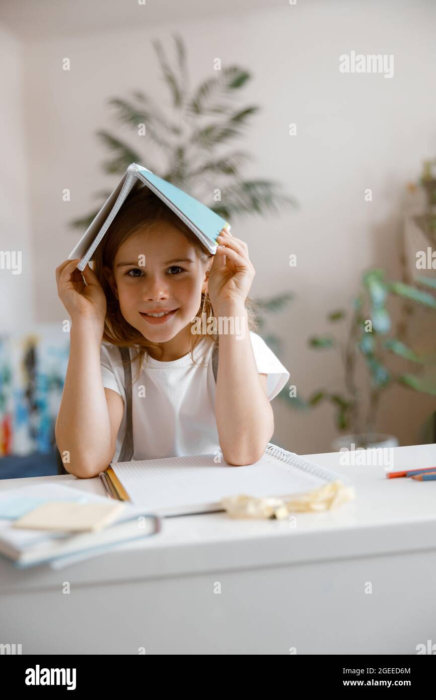 La ragazza piccola felice tiene il taccuino sulla testa seduta al tavolo nella stanza luminosa Foto Stock