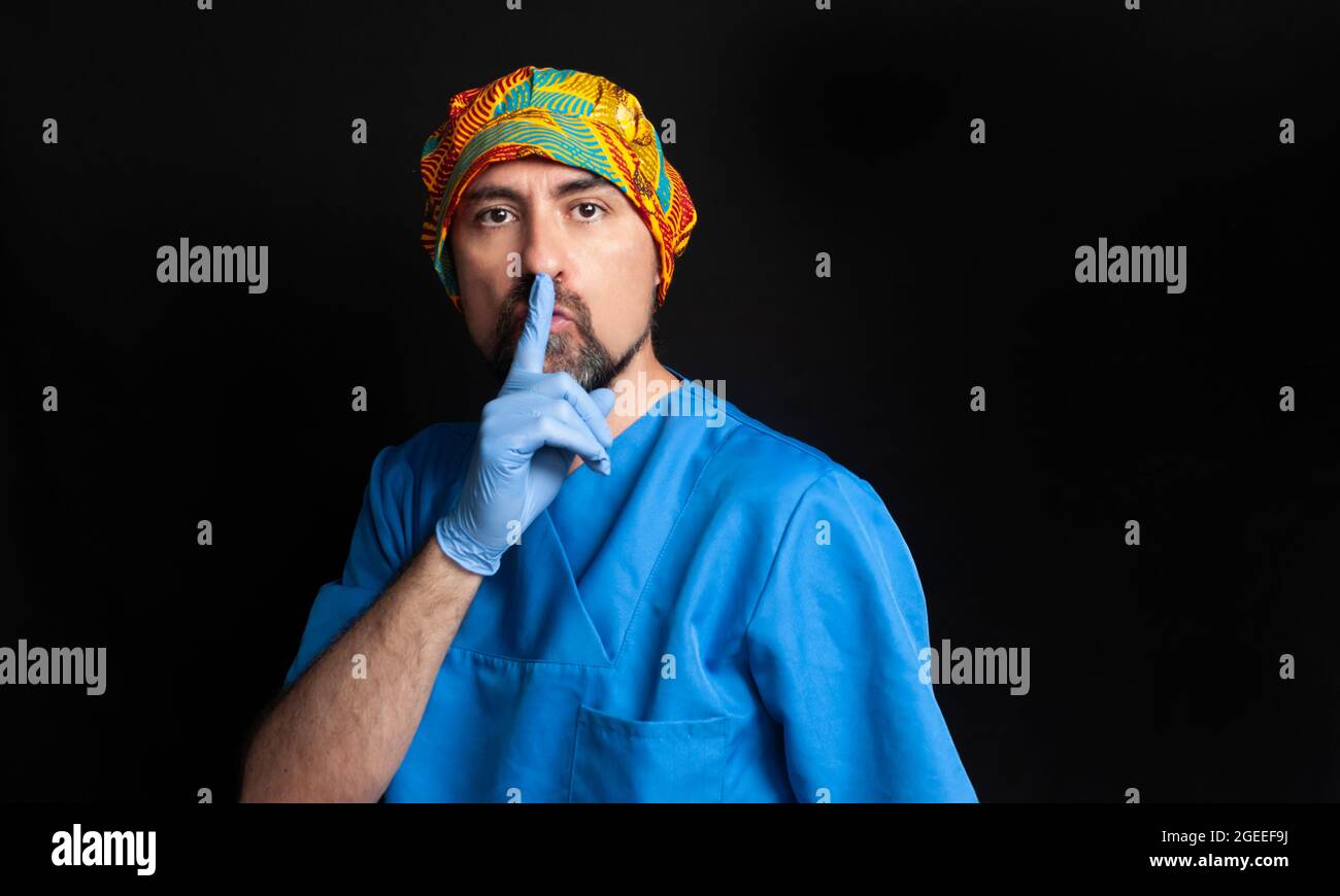 Un medico vestito in una divisa blu dell'ospedale, che indossa un cappuccio chirurgico etnico, scuro e bearded, chiede il silenzio con il dito indice sopra le labbra. R Foto Stock