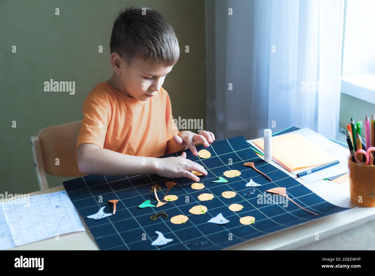 Cute bambino che crea Halloween-tema gioco da tavolo seduta da scrivania.  Ragazzo arrangiando elementi di gioco carta-tagliati sul campo di gioco  nero. Idee fai da te per i bambini Foto stock 