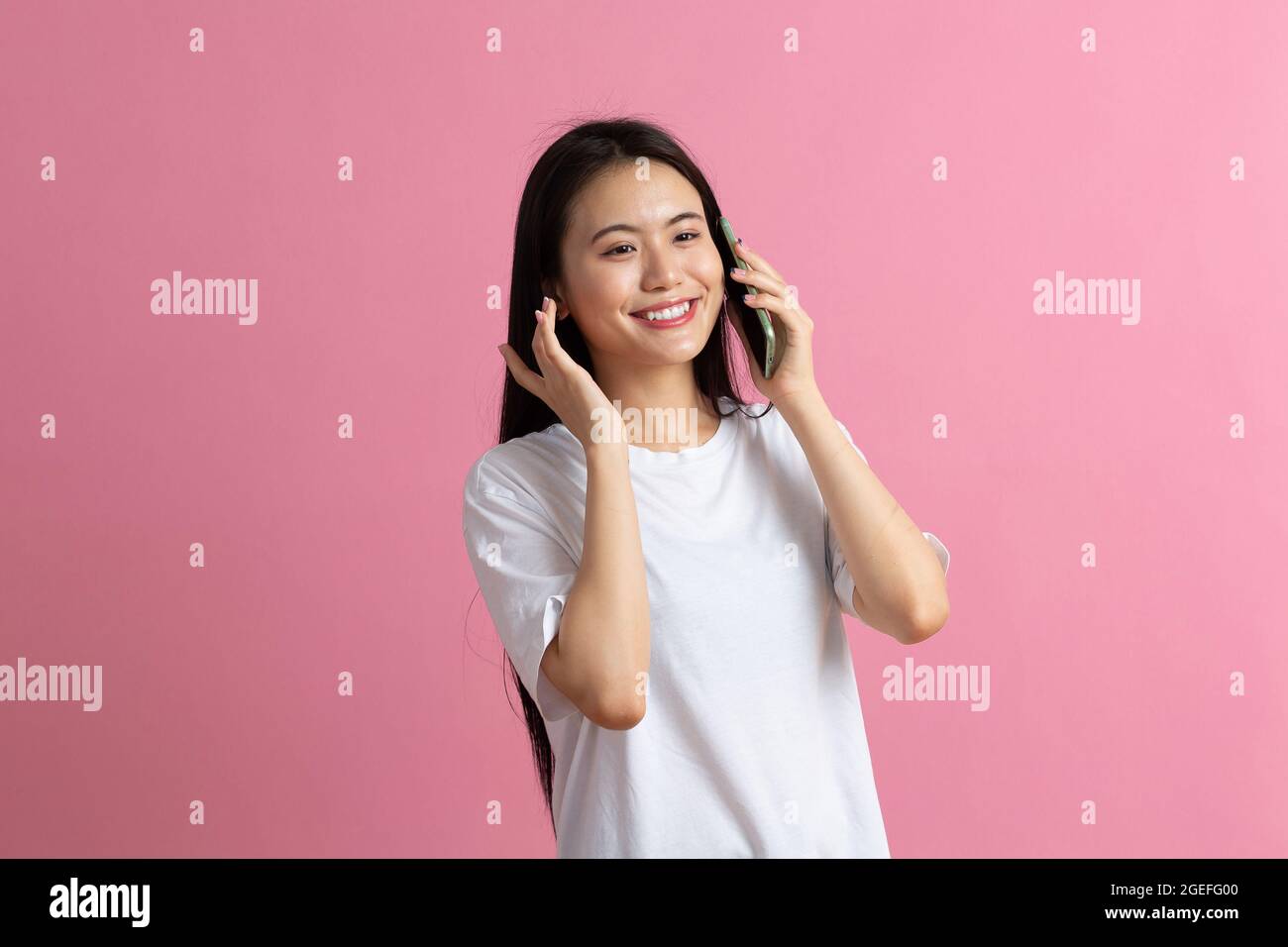 Donna asiatica sorridente che parla il telefono cellulare sull'orecchio in piedi su sfondo rosa Foto Stock