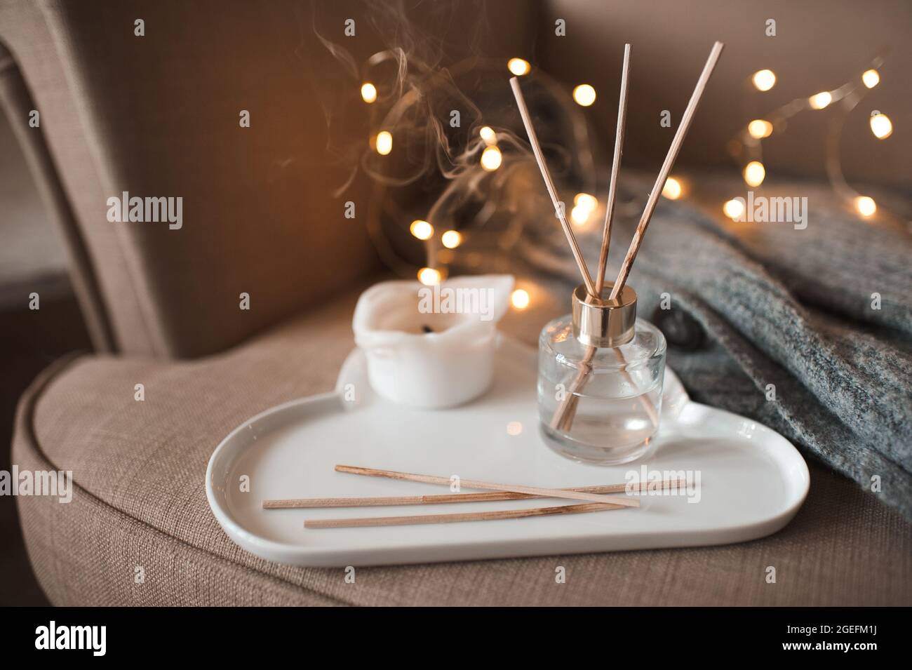 Profumo domestico liquido in bottiglia di vetro con bastoni di bambù di legno e candela bruciante su vassoio di ceramica bianca, cardigan di lana lavorato a maglia in una comoda sedia in camera Foto Stock