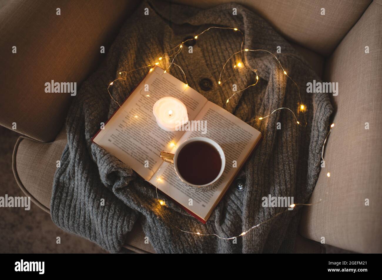 Tazza di tè nero fresco soggiorno sul libro di carta aperta con maglione di lana a maglia in sedia accogliente sopra le luci di Natale incandescente primo piano. Vista dall'alto. Colazione di prima mattina Foto Stock