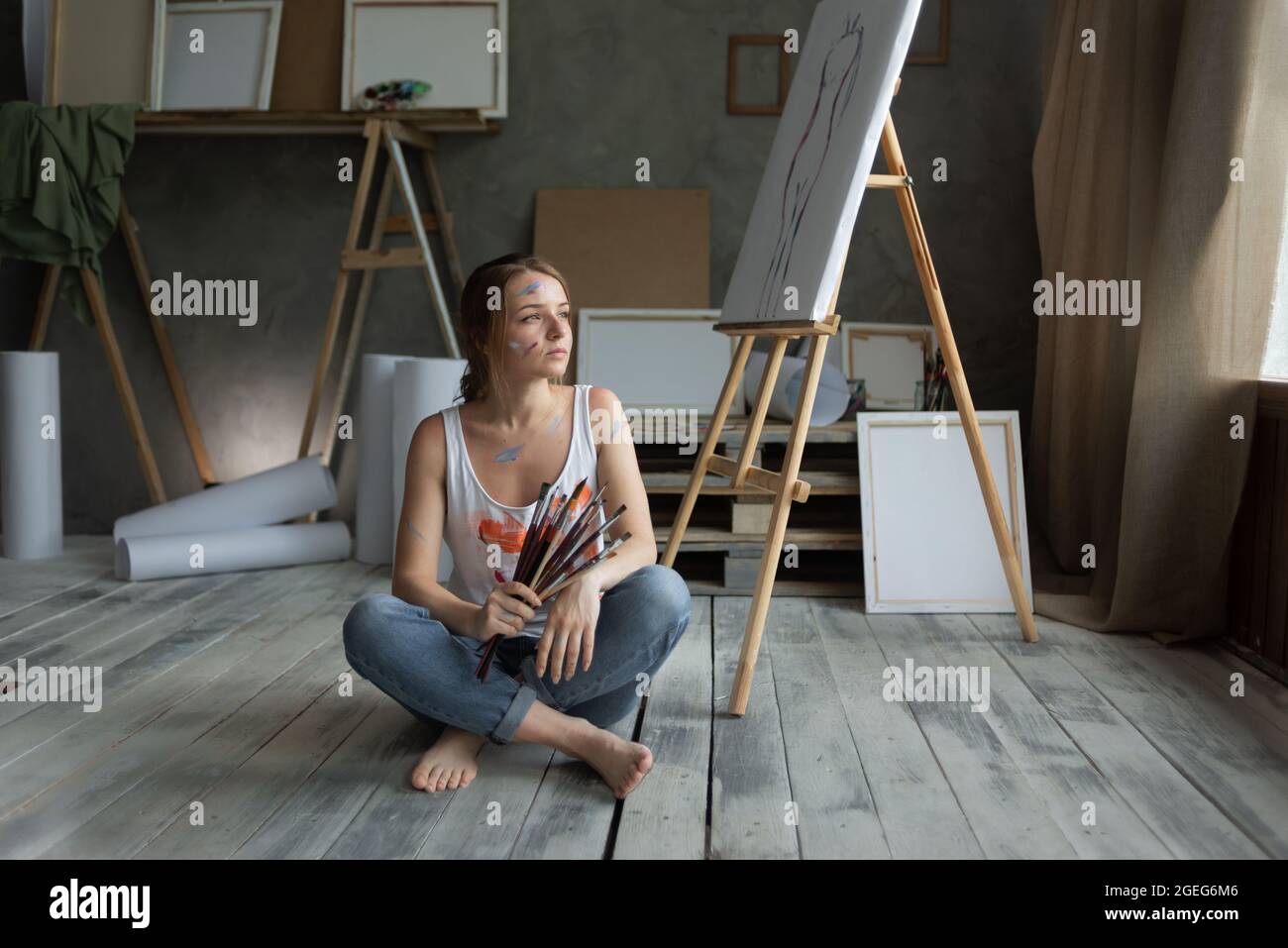 una giovane bella donna artista siede sul pavimento in uno studio d'arte che tiene un pennello nelle sue mani. Foto Stock