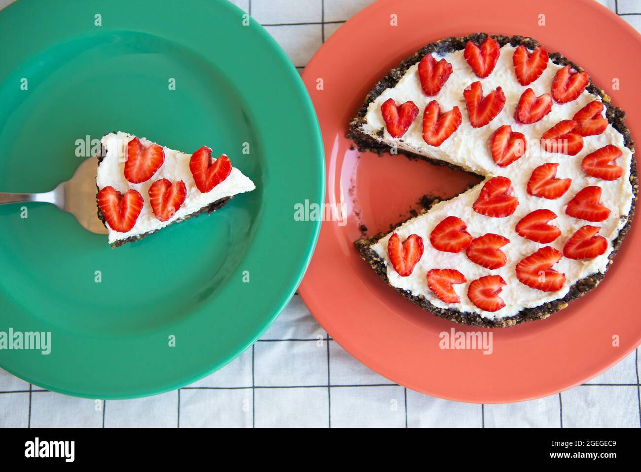 Una cheesecake alla fragola decorata con fragole a forma di cuore si trova  su un piatto e si trova su un tovagliolo a scacchi, tagliare una fetta.  Vista dall'alto Foto stock -