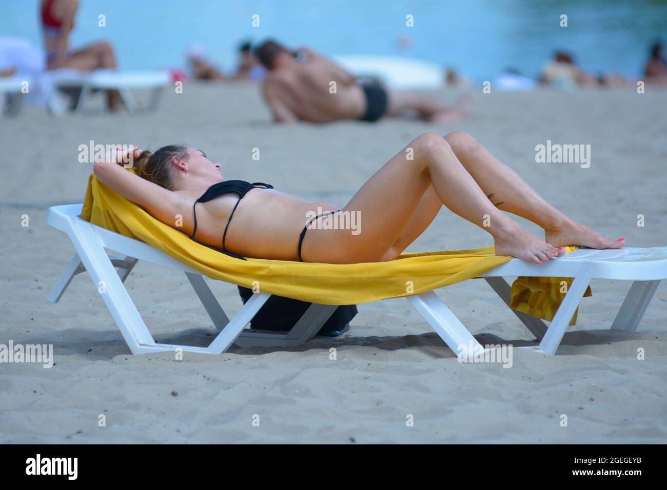 Giovane donna sdraiata sulla spiaggia chaise lounge prendere il sole su una spiaggia di sabbia fiume, offuscato persone su uno sfondo. 2 agosto 2012. Kiev, Ucraina Foto Stock