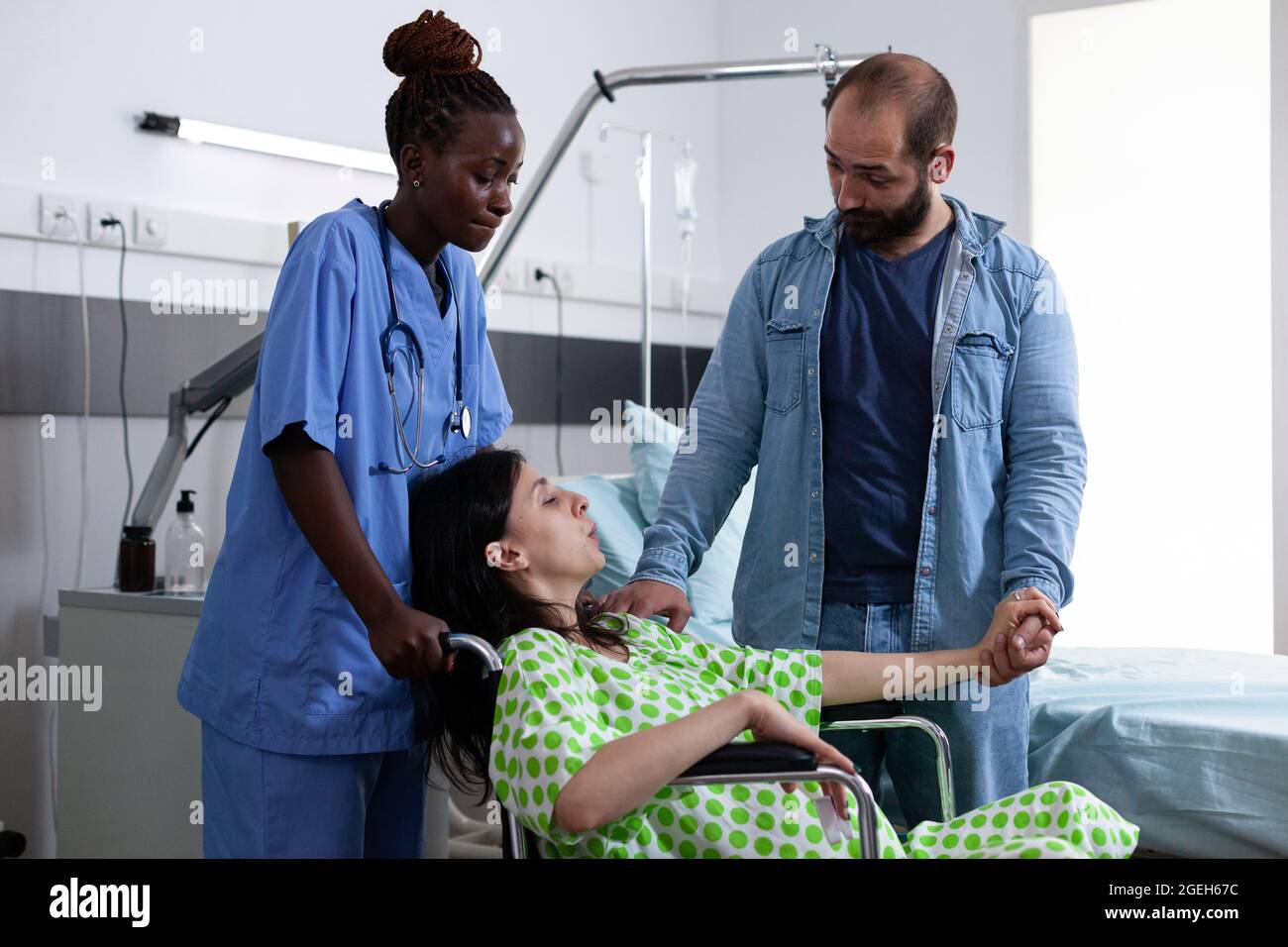Donna incinta in sedia a rotelle ricevere assistenza da infermiera e marito mentre si prepara per il parto nella stanza di maternità in clinica. Assistente medico che aiuta la coppia che aspetta il bambino Foto Stock