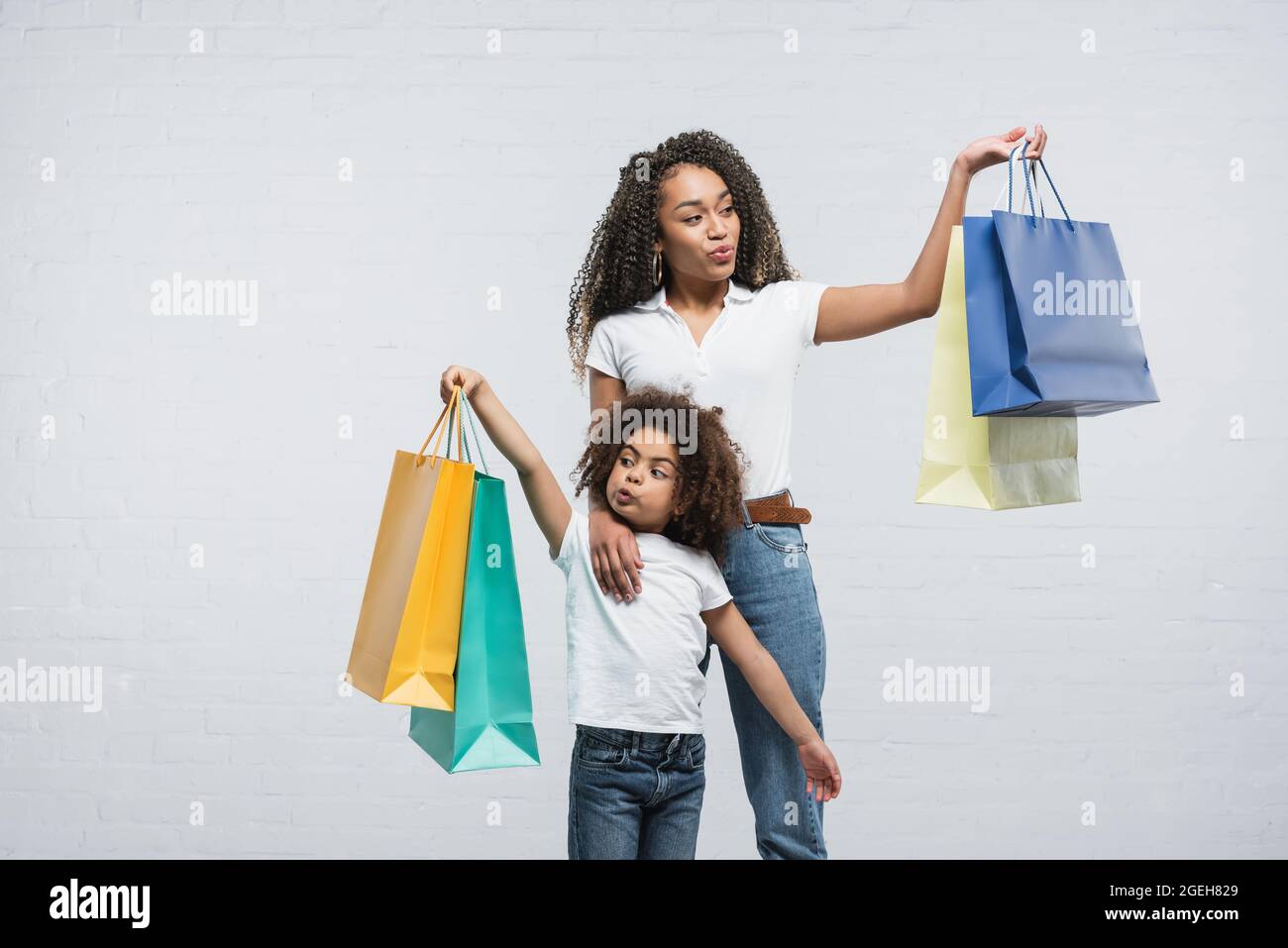 stupita donna afroamericana con la figlia puffing fuori guance mentre tiene i sacchetti di shopping sul grigio Foto Stock