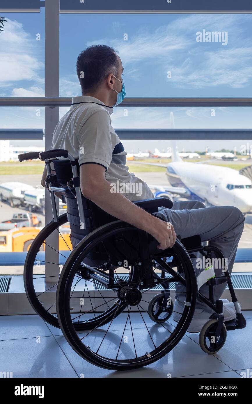 Giovane uomo con maschera protettiva in sedia a rotelle all'aeroporto guardando fuori dalla finestra Foto Stock