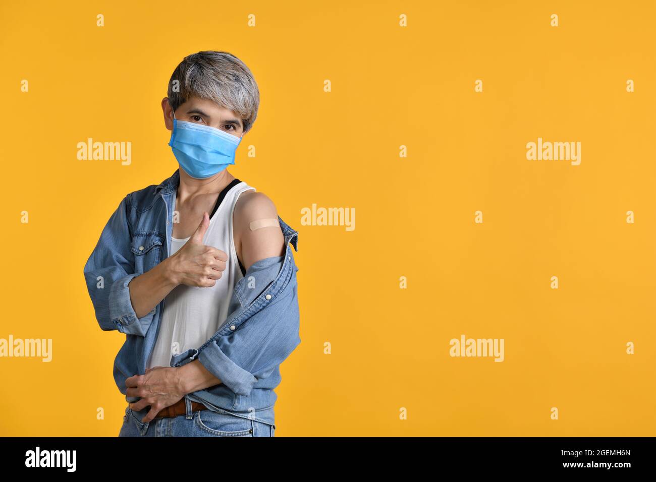 Donna asiatica di mezza età felice vaccinata mostrando i pollici in su a cerotto adesivo sul suo braccio dopo l'iniezione antivirale di coronavirus isolato su YE Foto Stock