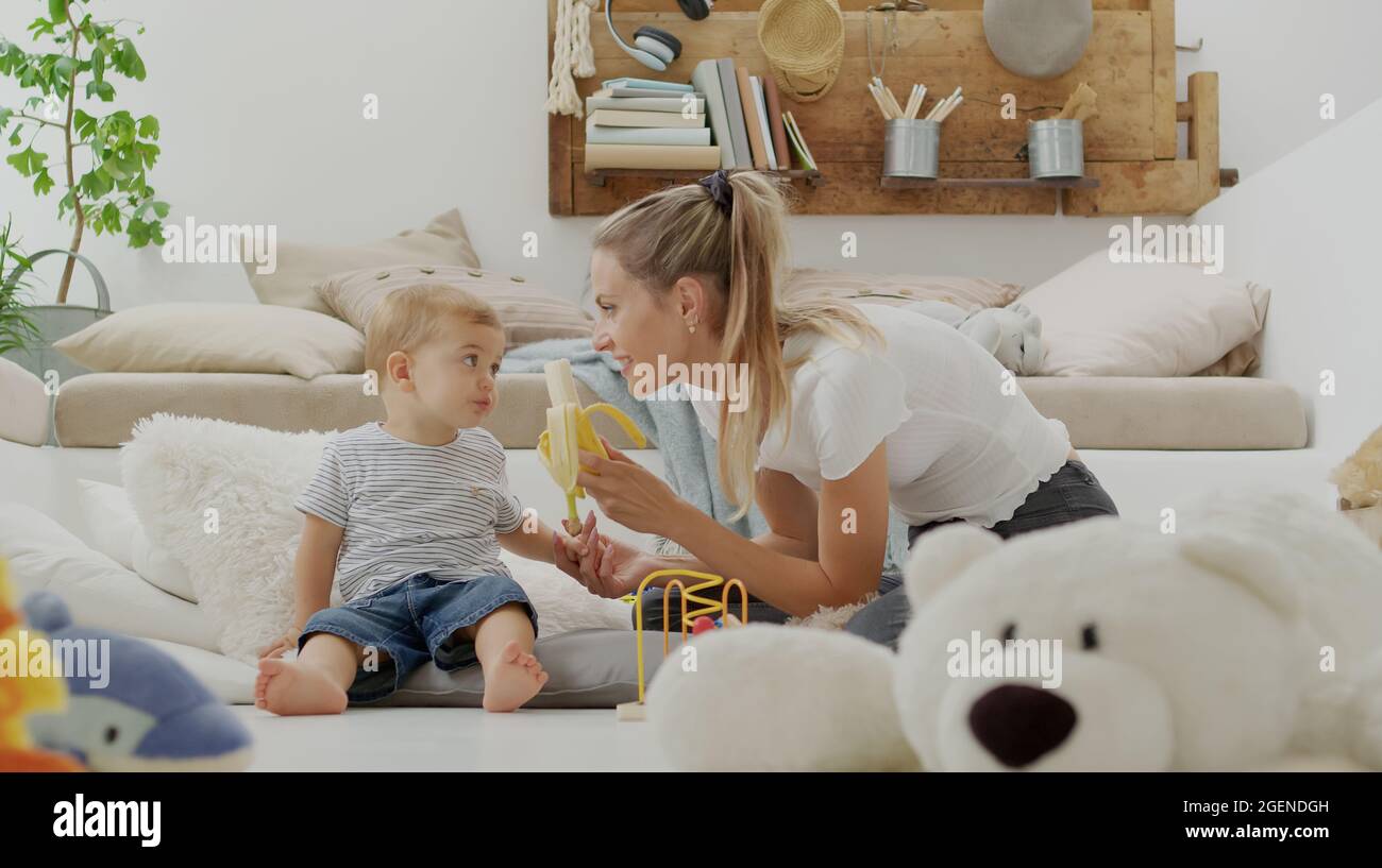 La mamma felice e sorridente rende il vostro bambino bello mangiare la banana per una dieta equilibrata ricca di vitamine e minerali, seduta a casa tra cuscini e Foto Stock