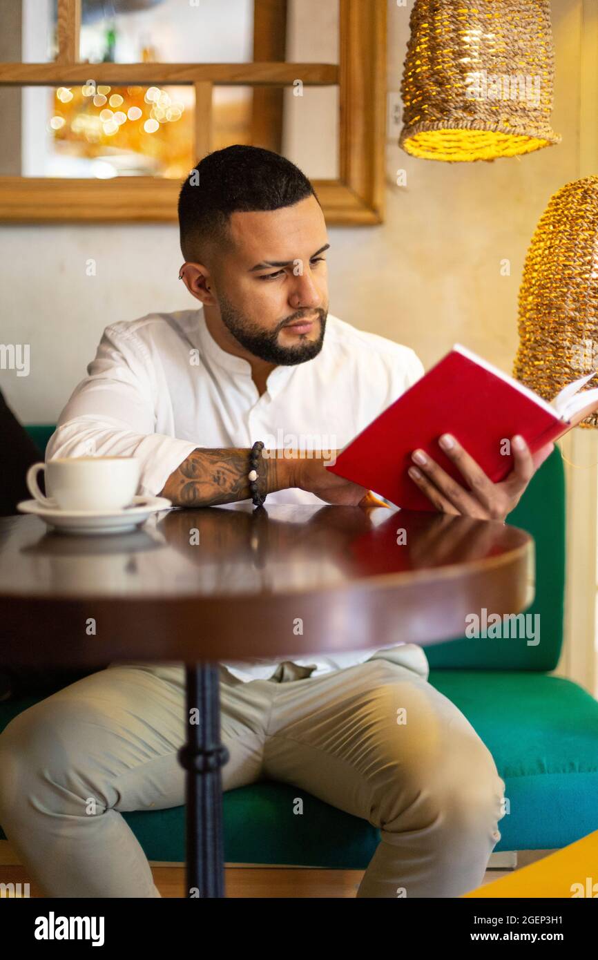 Ritratto di giovane ragazzo latino che si gode di leggere un libro in una moderna caffetteria. Foto Stock