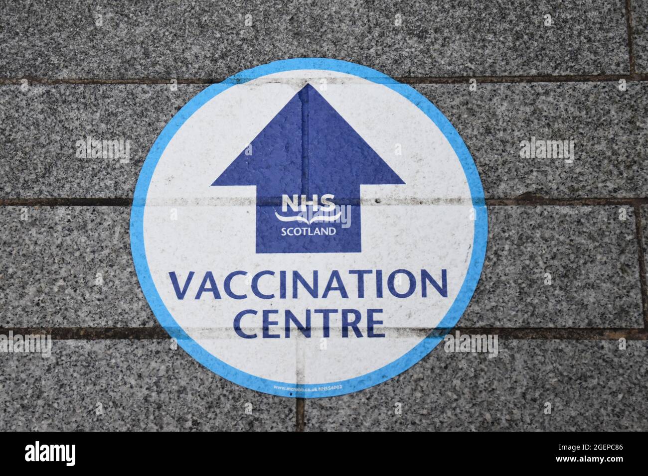 Segno isolato NHS Scotland Vaccine Center in cerchio blu con freccia. Su marciapiede con pietre grigie. Foto Stock