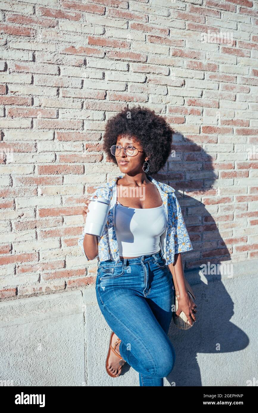 giovane afroamericana in piedi appoggiata al muro. Sta tenendo una tazza bianca riutilizzabile di caffè. Foto Stock