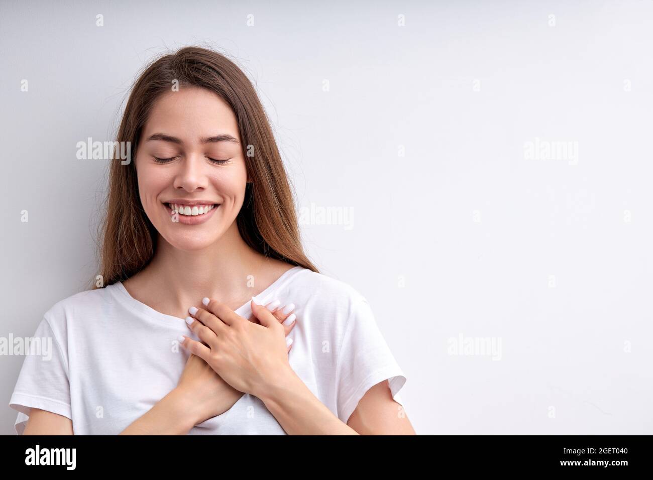 Donna piuttosto felice esprimente gratitudine, tenendo le mani sul petto, grato per tutto, con gli occhi chiusi, con un bel sorriso, vestito casualmente. isola Foto Stock